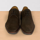 A pair of dark brown suede lace-up shoes with speckled soles, displayed on a light wood surface against a white background.