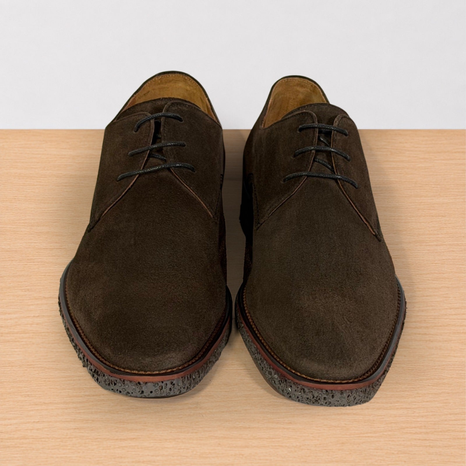 A pair of dark brown suede lace-up shoes with speckled soles, displayed on a light wood surface against a white background.