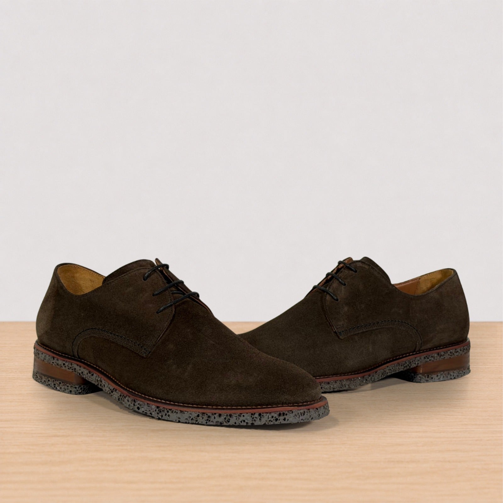 A pair of dark brown suede derby shoes with laces and speckled soles, displayed on a light wood surface against a plain light gray background.