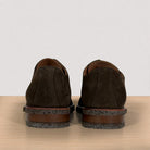 A pair of dark brown suede shoes shown from the back, resting on a light wood surface against a plain off-white background.