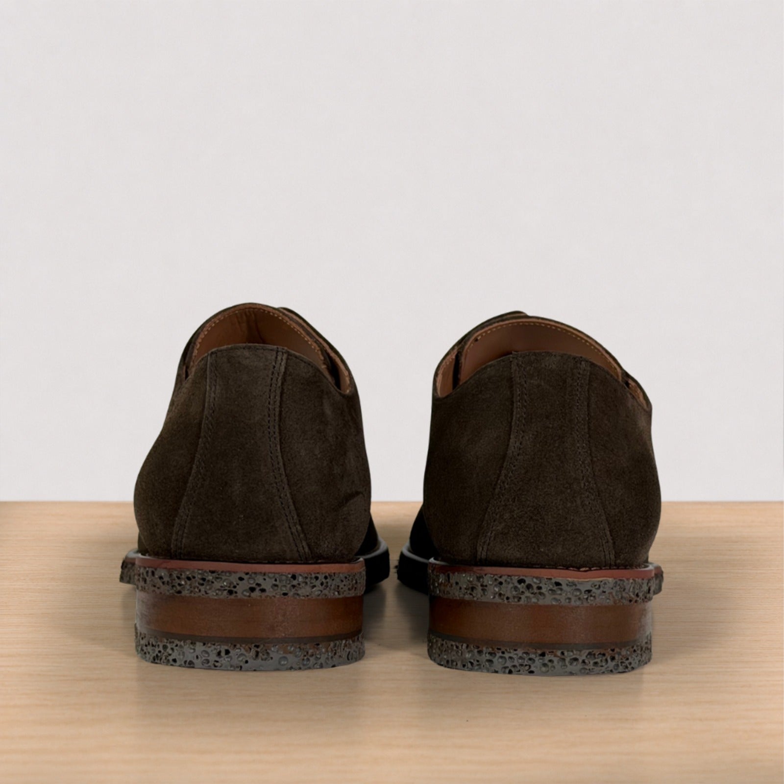 A pair of dark brown suede shoes shown from the back, resting on a light wood surface against a plain off-white background.
