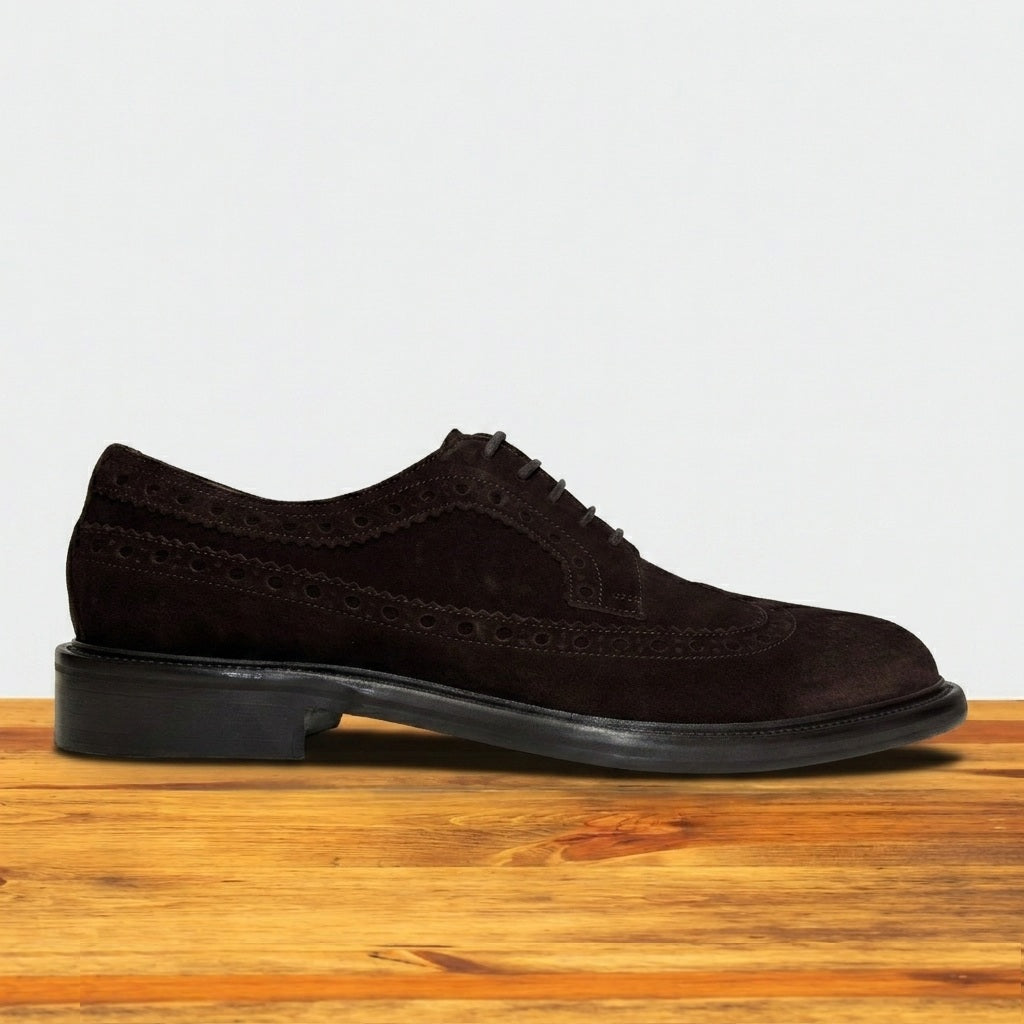Chocolate brown suede wingtip shoe with decorative perforations and lace-up closure, displayed on a rustic wooden surface against a light grey background
