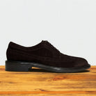 Chocolate brown suede wingtip shoe with decorative perforations and lace-up closure, displayed on a rustic wooden surface against a light grey background