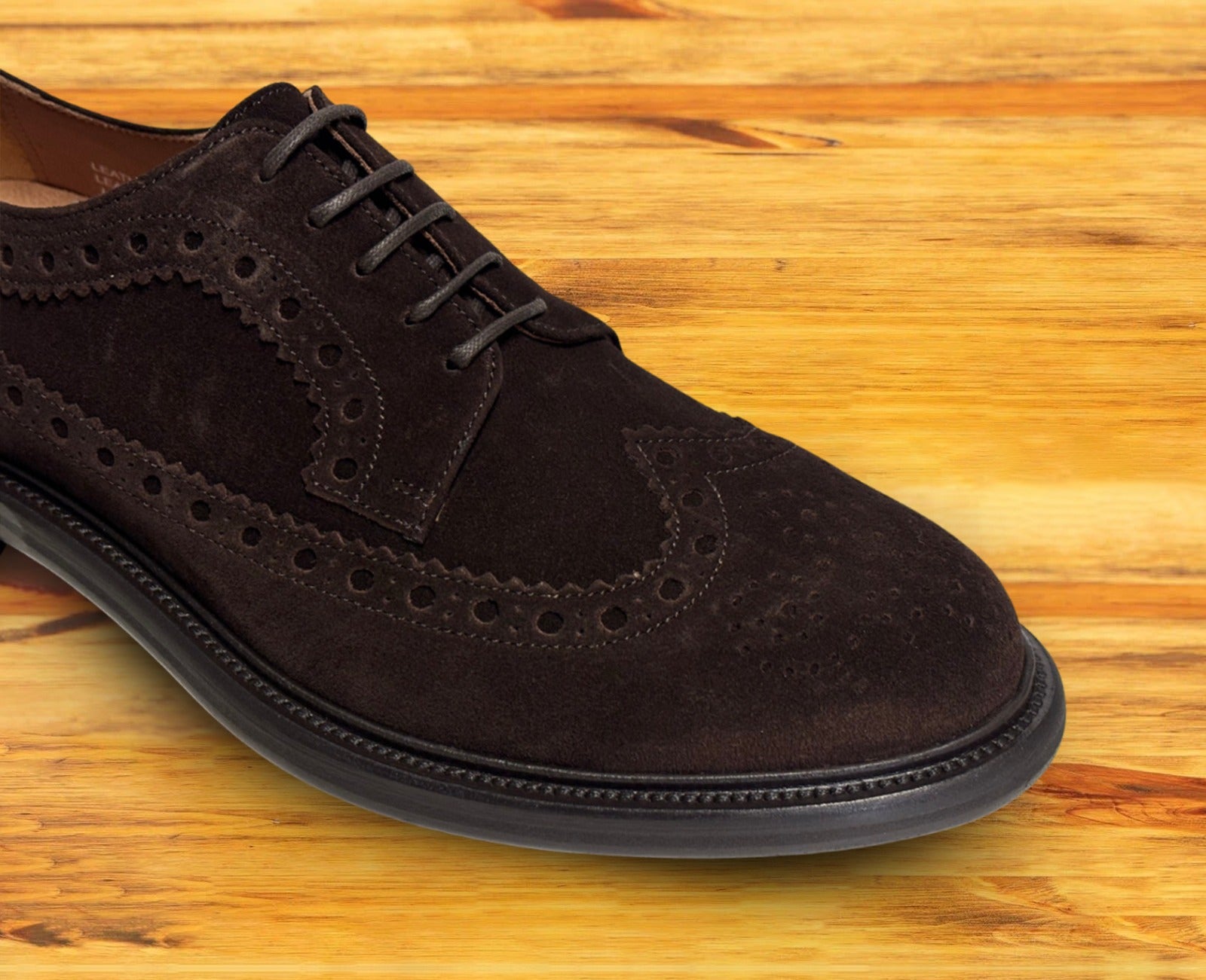 Close view of Chocolate brown suede wingtip shoe with decorative perforations and lace-up closure, displayed on a wooden surface