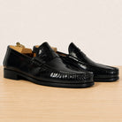 A pair of black leather penny loafers with a glossy finish, displayed on a light wood surface against a warm beige background.