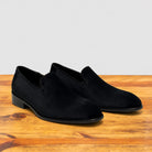 A pair of black suede slip-on loafers with low block heels, displayed on a rustic wooden surface against a light gray background.