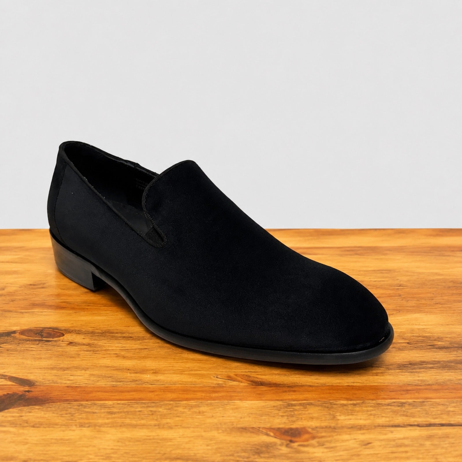 A single black suede slip-on loafer with a low block heel, displayed on a wooden surface against a light gray background.