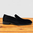 A single black suede slip-on loafer shoe with a low block heel, displayed on a wooden surface against a light gray background.