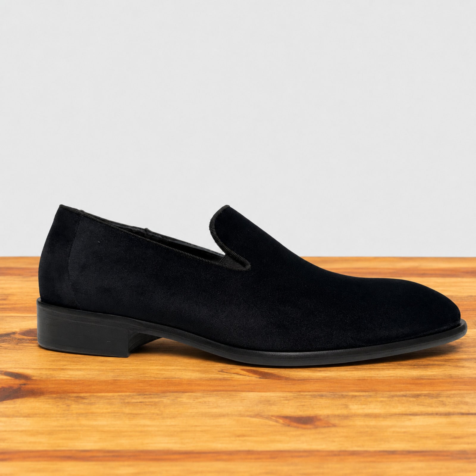 A single black suede slip-on loafer shoe with a low block heel, displayed on a wooden surface against a light gray background.