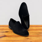 A pair of black suede slip-on loafers displayed on a wooden surface against a light gray background.