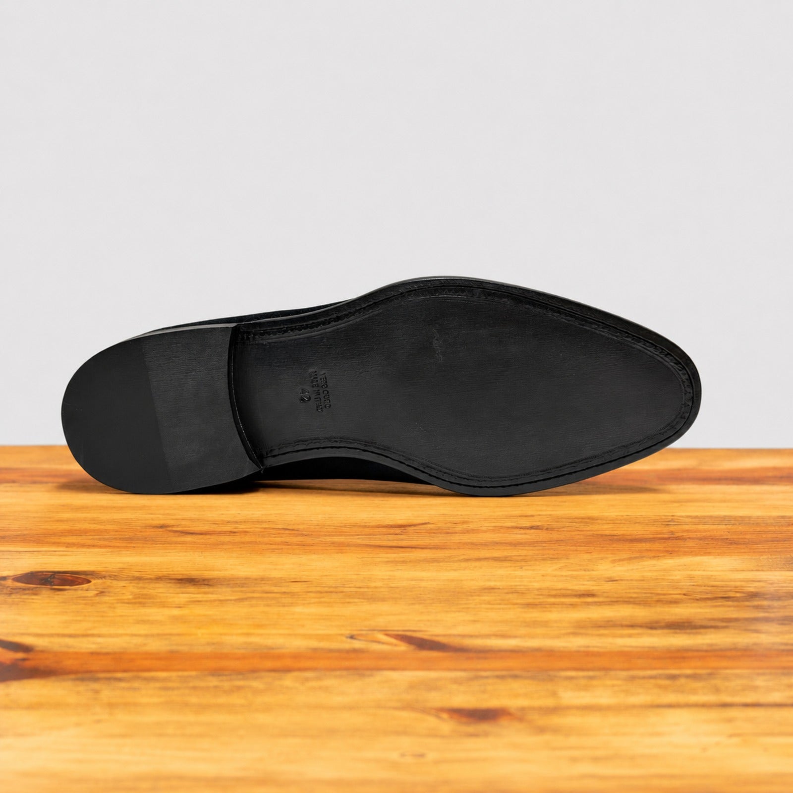 The black outsole of a dress shoe is shown from a top-down view, resting on a wooden surface against a light gray background.