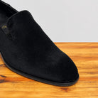 A close-up of a black suede slip-on shoe resting on a wooden surface against a light gray background.
