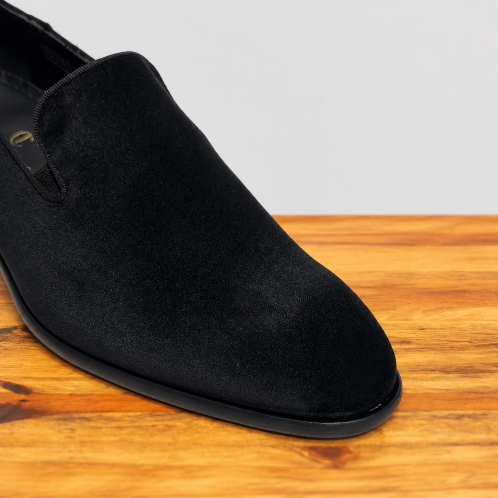 A close-up of a black suede slip-on shoe resting on a wooden surface against a light gray background.