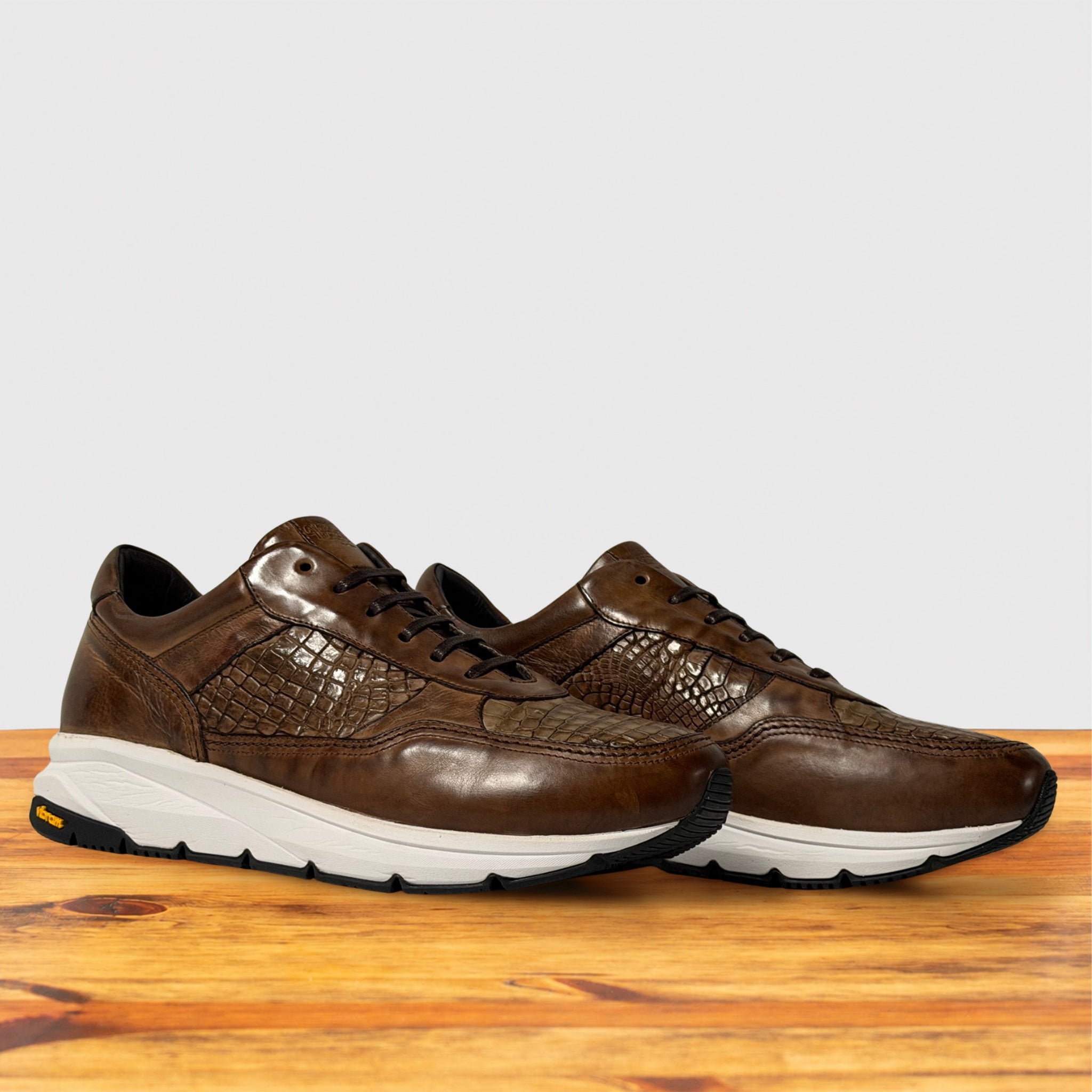 A pair of moor color leather athletic sneakers with texture and thick white midsoles, displayed on a wooden surface against a light gray background.
