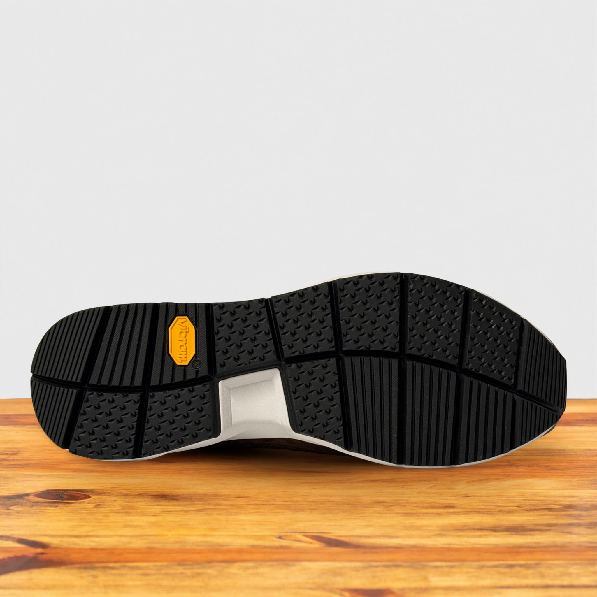 The black rubber outsole of a shoe resting on a wooden surface against a light gray background. A small yellow Vibram logo is visible on the rear section of the tread.