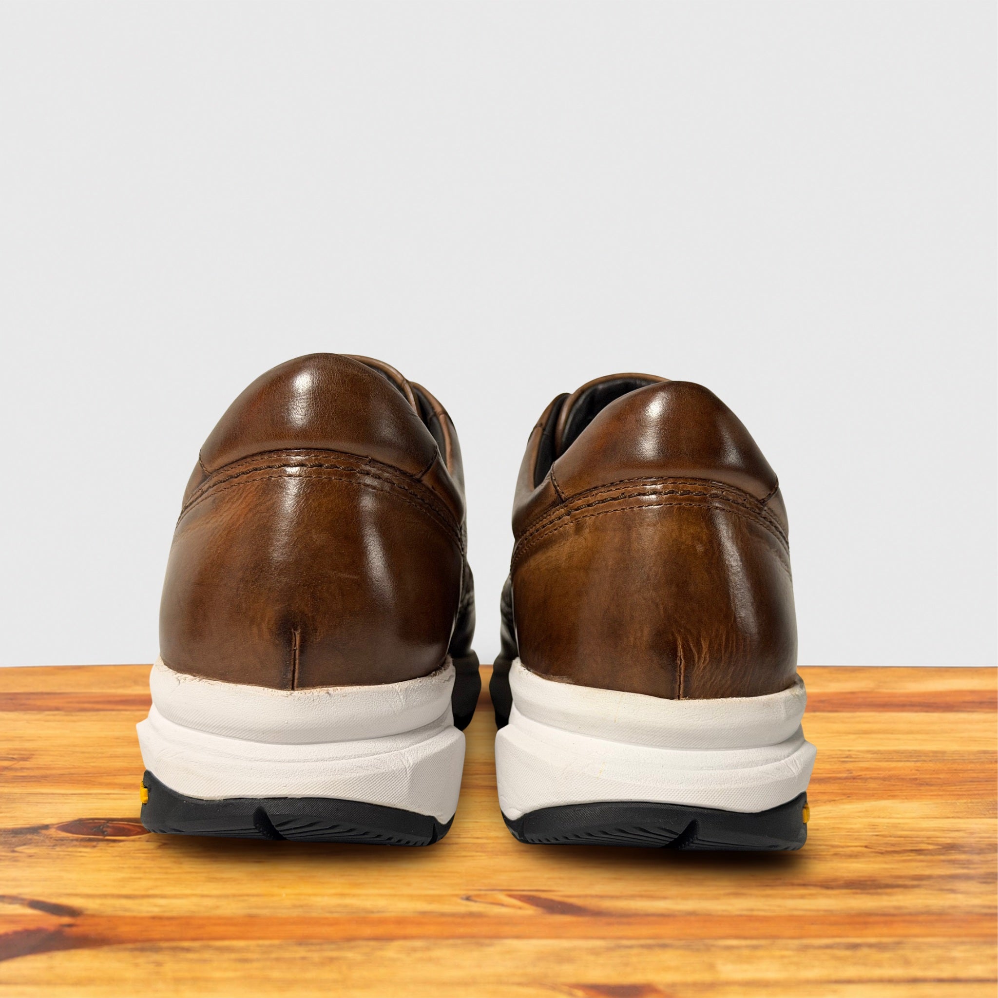 A pair of brown leather dress shoes viewed from the back, resting on a wooden surface against a light gray background.