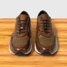 A pair of brown leather low-top sneakers with textured crocodile-pattern panels, shown from the front on a wooden surface against a light gray background.