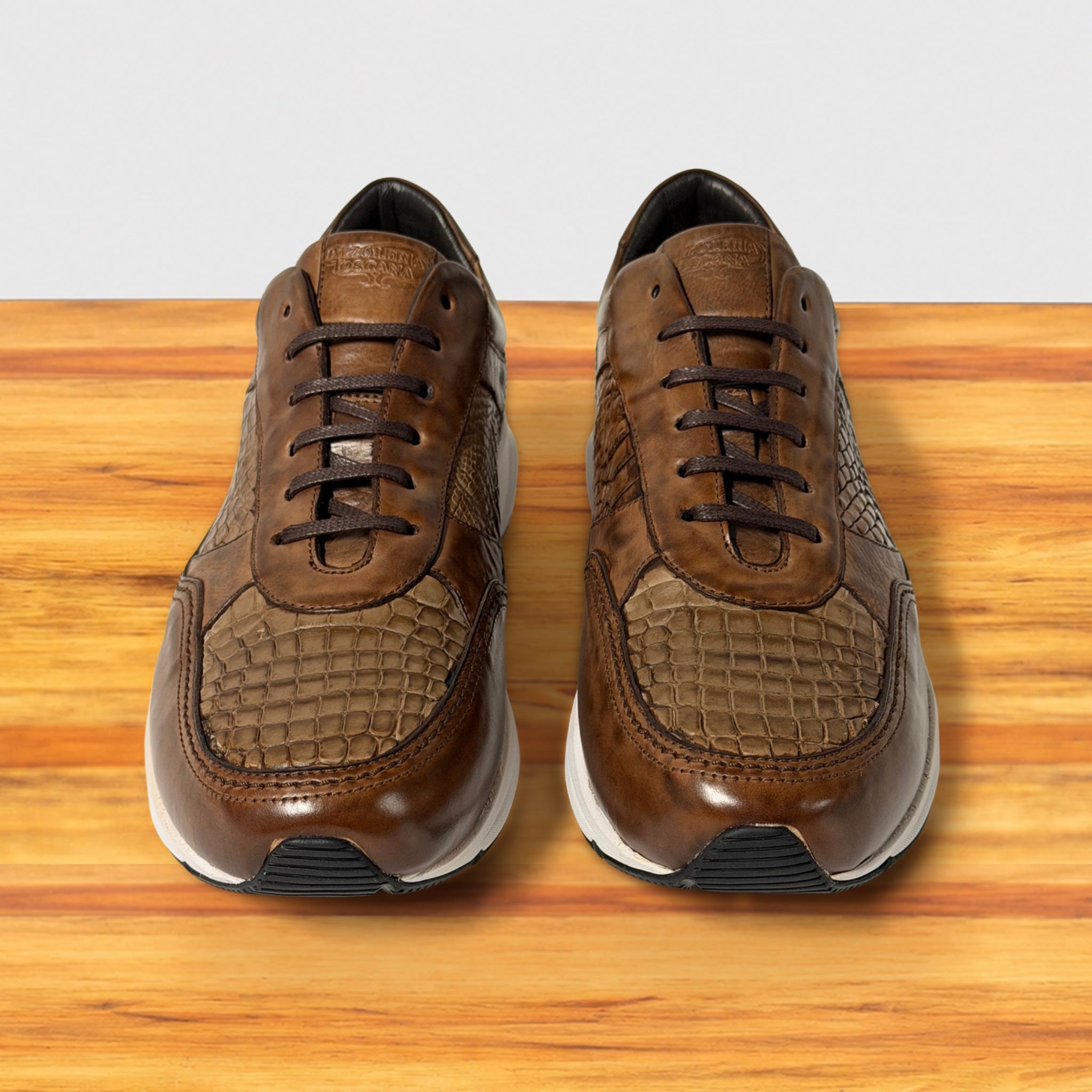 A pair of brown leather low-top sneakers with textured crocodile-pattern panels, shown from the front on a wooden surface against a light gray background.