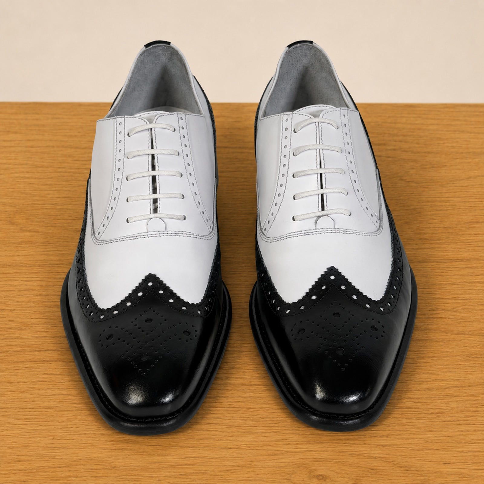 A pair of men's wingtip oxford shoes in a two-tone black and white colorway, displayed from a top-down view on a light wood surface.