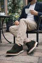 Person sitting outdoors holding a cup, wearing a Olive Green suede wingtip derby shoe with a rubber sole.