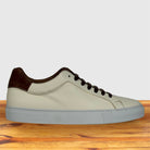 A low-top sneaker with a light beige leather upper, dark brown heel tab, brown laces, and a light gray sole, displayed on a wooden surface against a light gray background.