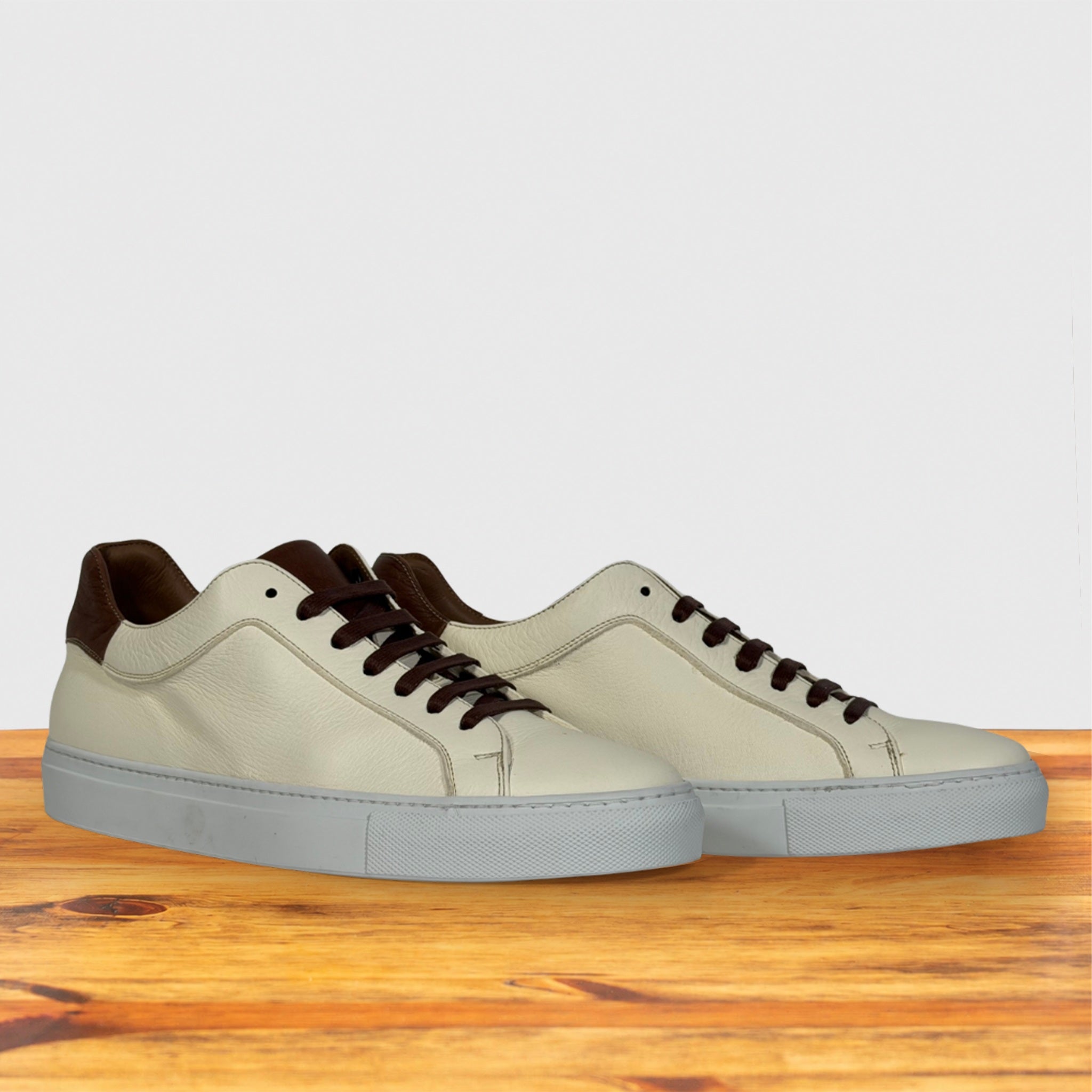 A pair of low-top leather sneakers in off-white with brown accents on the collar, tongue, and laces, resting on a wooden surface against a light gray background.