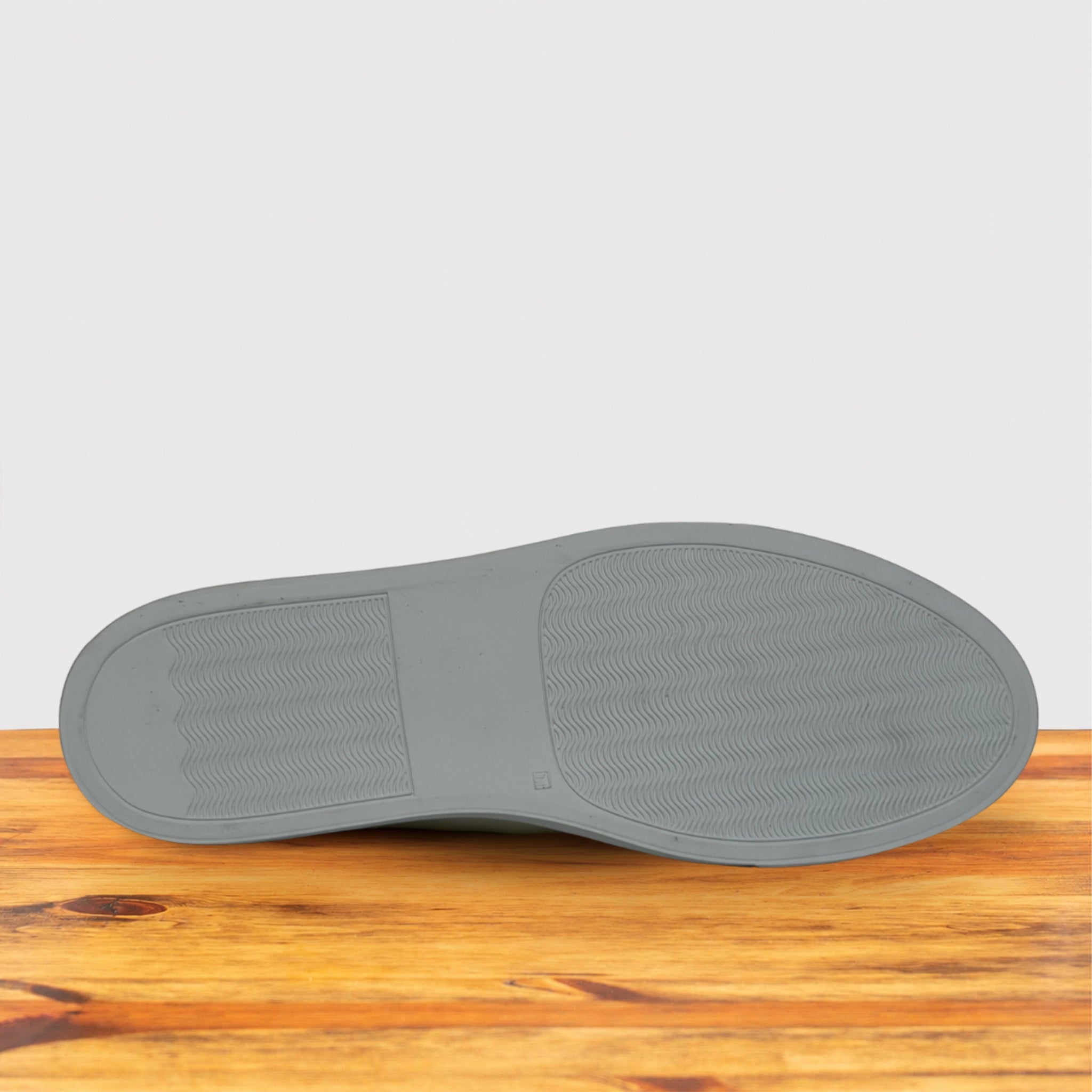 The outsole of a shoe shown from a bottom-up perspective, resting on a wooden surface against a light gray background.