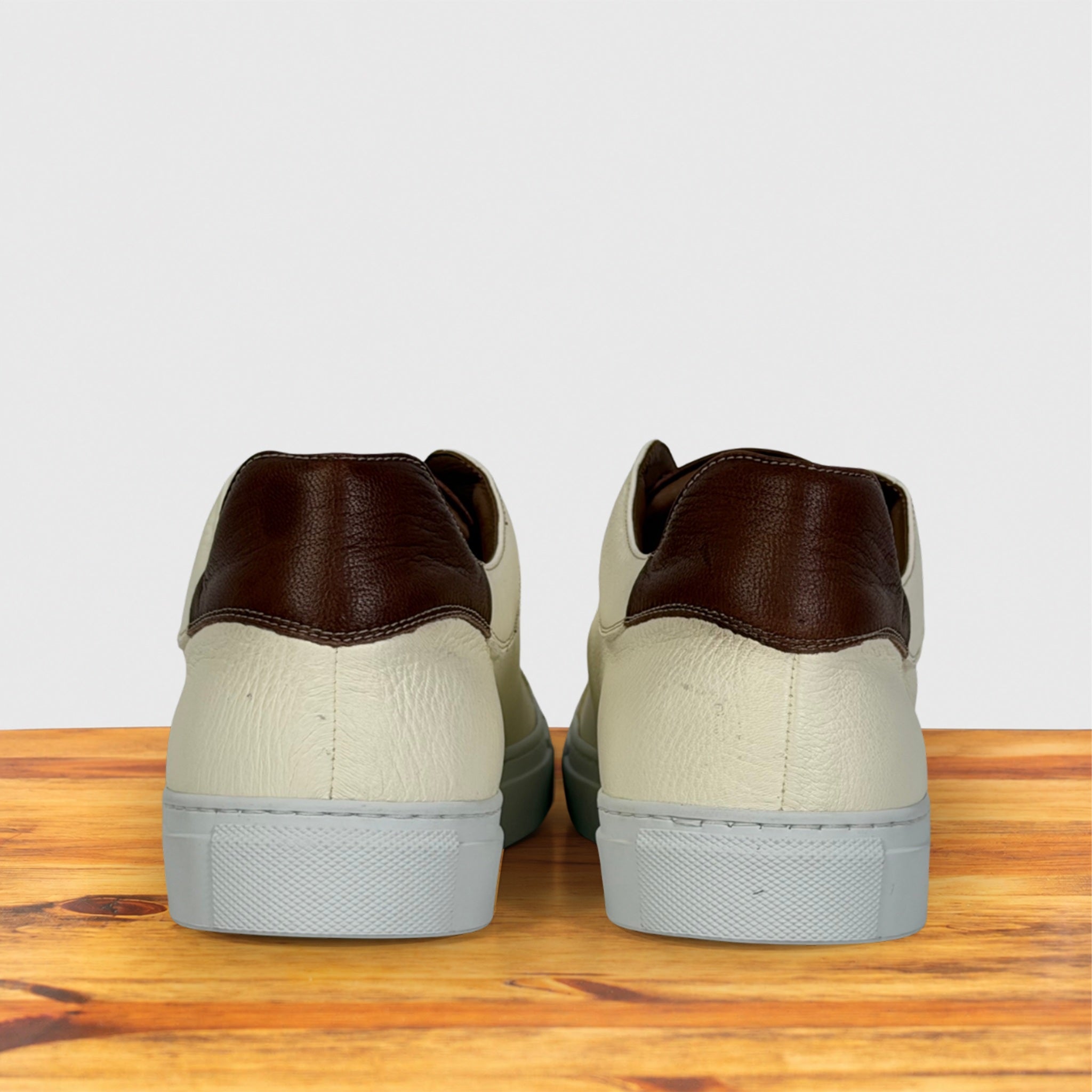 A pair of low-top sneakers shown from the back, featuring dark brown leather heel panels, textured cream leather uppers, and light gray rubber soles, displayed on a wooden surface against a light gray background.