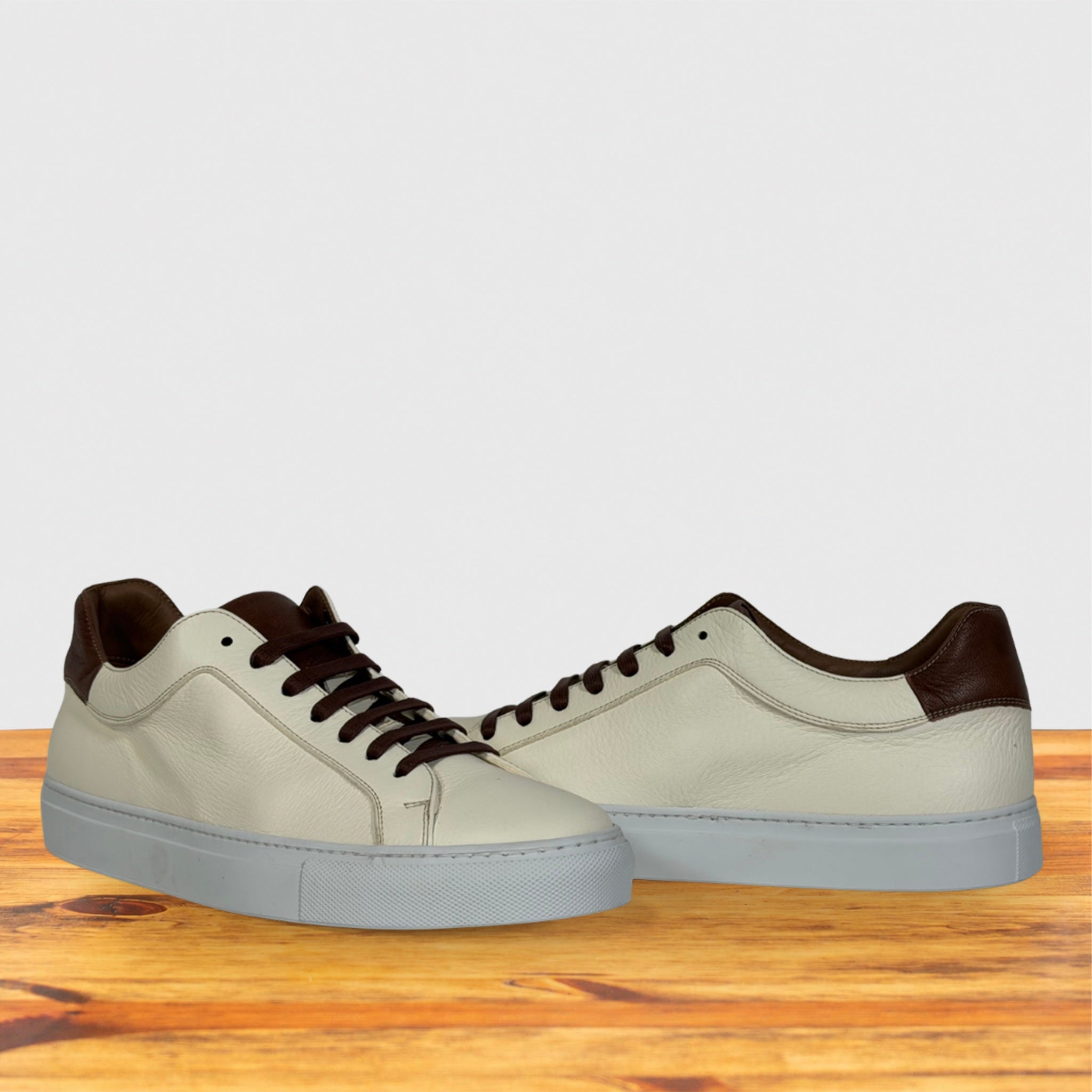 A pair of low-top sneakers with cream leather uppers, brown laces, displayed on a wooden surface against a light gray background.
