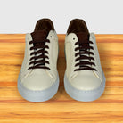A pair of low-top sneakers shown from the front on a wooden surface with a light gray background. The shoes have cream leather uppers, dark brown laces, brown interiors