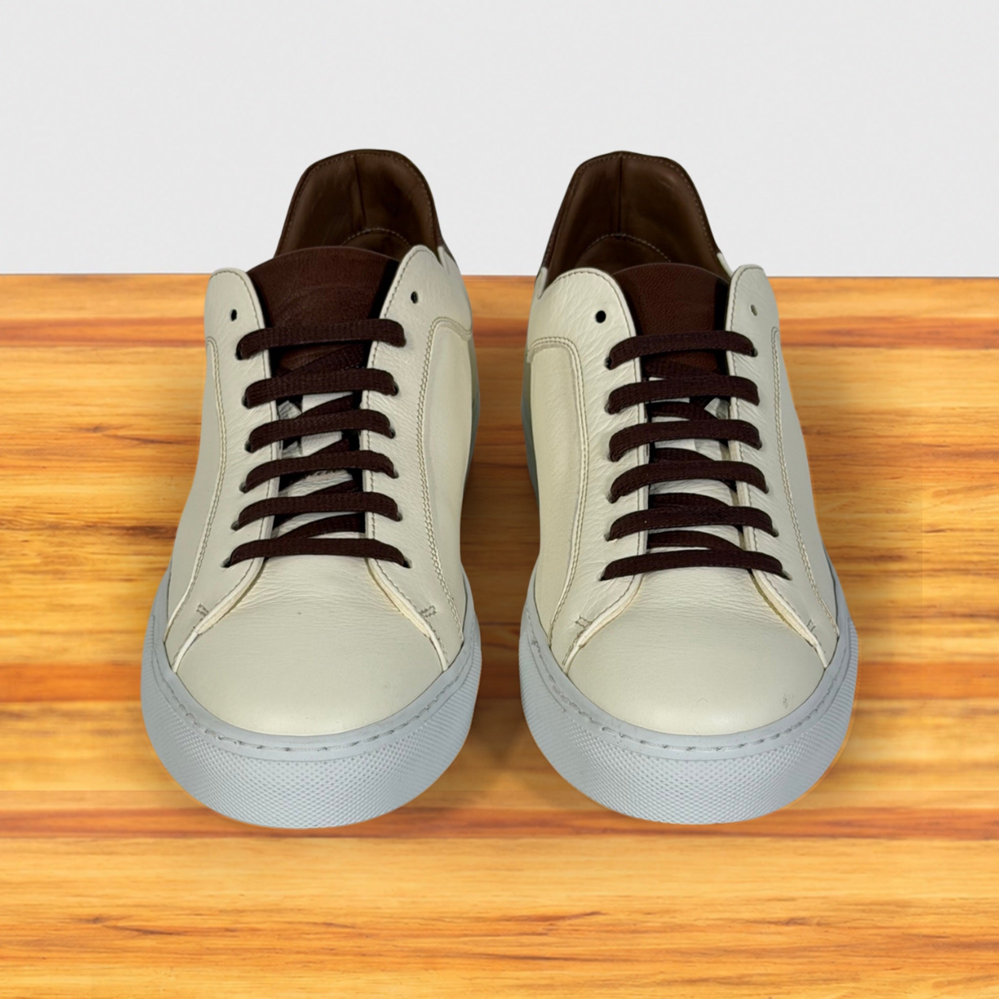 A pair of low-top sneakers shown from the front on a wooden surface with a light gray background. The shoes have cream leather uppers, dark brown laces, brown interiors