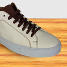 A low-top sneaker with a light cream leather upper, dark brown laces and collar,  shown on a wooden surface against a white background.