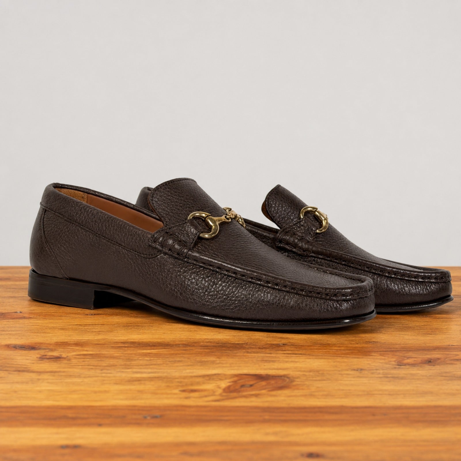 A pair of dark brown leather loafer with a gold buckle on a wooden surface