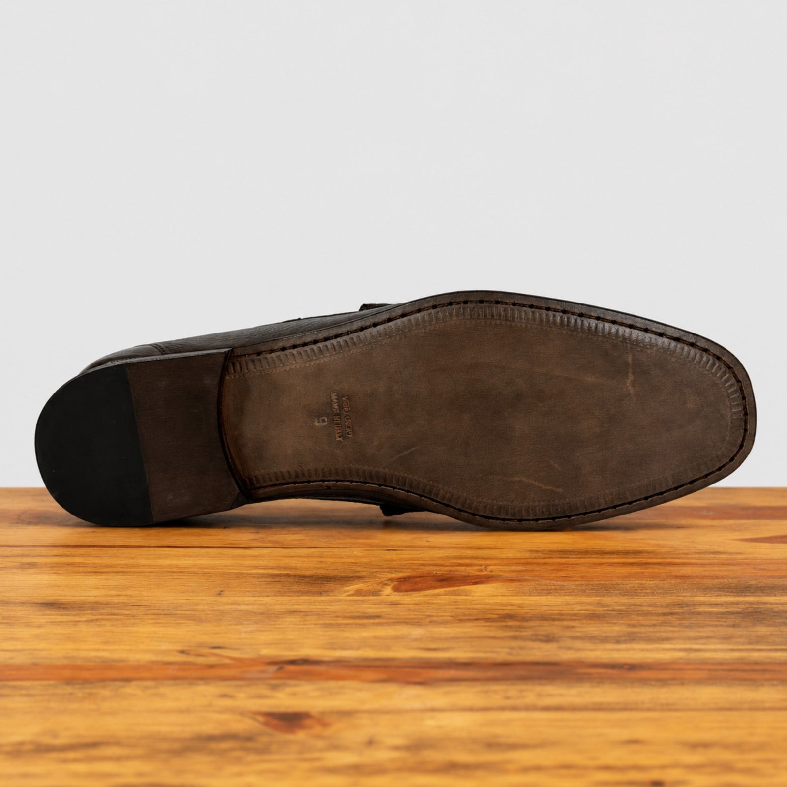 The sole of a dark brown leather dress shoe, showing the textured outsole and heel, resting on a wooden surface against a light gray background.
