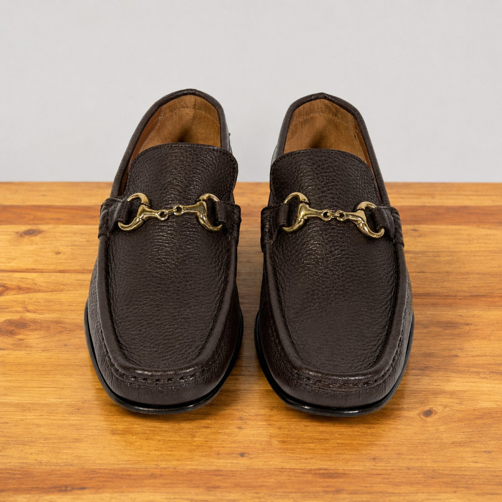 A pair of dark brown  leather penny loafers with gold buckle on a wooden surface against a neutral light gray background.