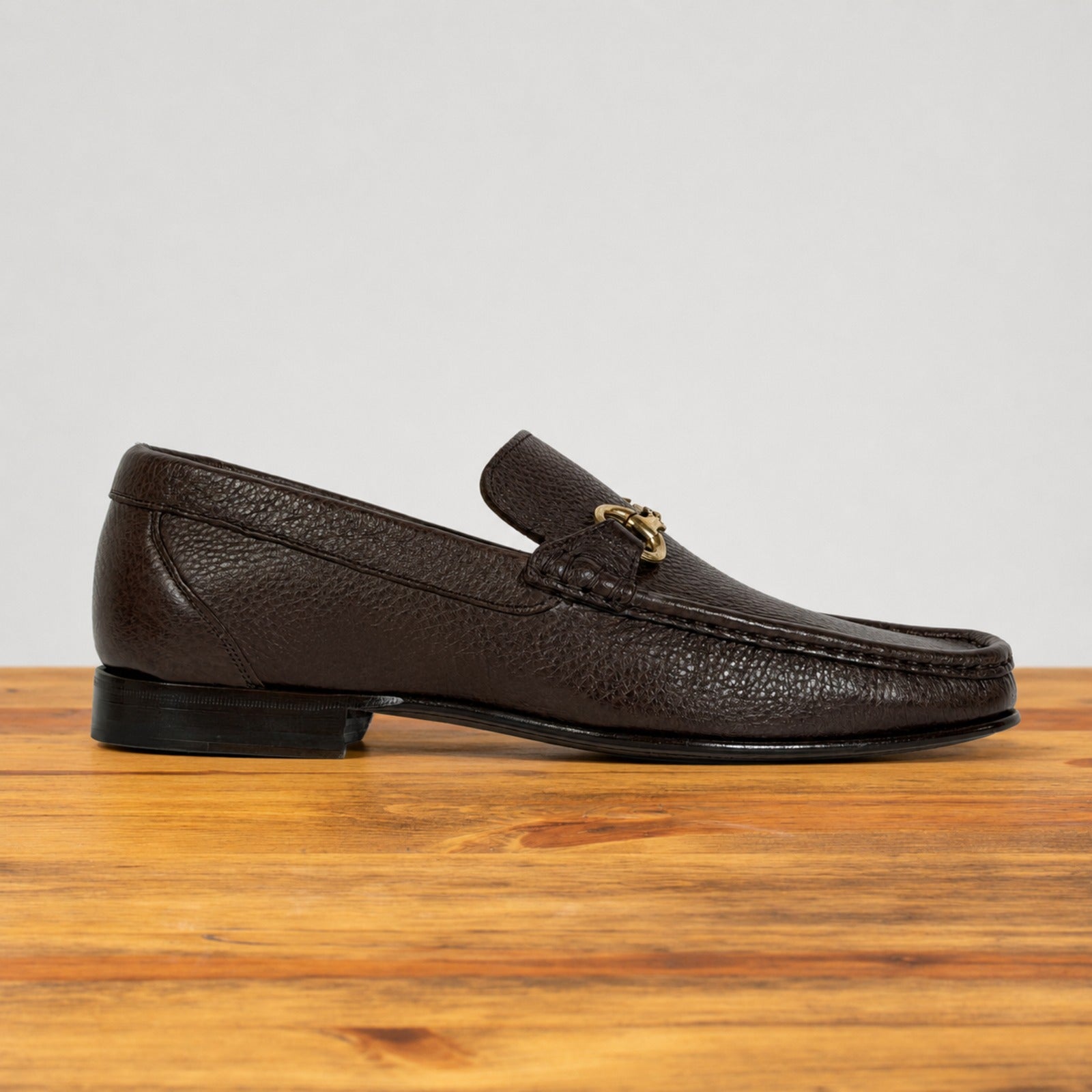 A dark brown  leather loafer with a gold-tone horsebit detail on the vamp, displayed in profile on a wooden surface against a light gray background.