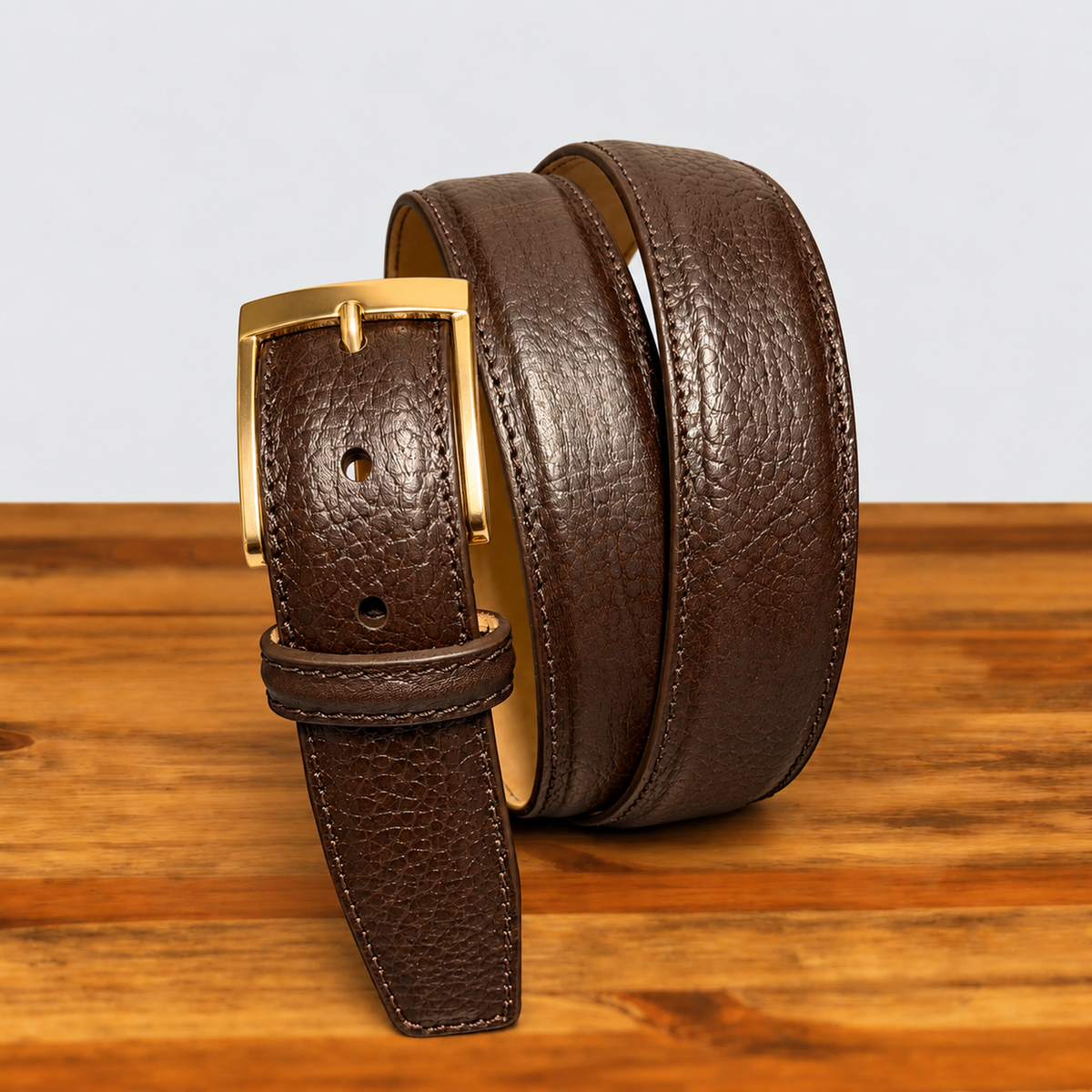 Dark brown textured leather belt with stitched edges and a gold-tone buckle, standing in loops on a wooden surface.
