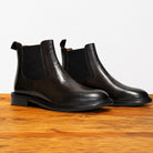 Pair of tobacco color leather Chelsea boots on a wooden surface with a light gray background