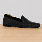 A single black leather loafer-style driving shoe with a textured pattern, shown in profile on a light wood surface against a neutral gray background.
