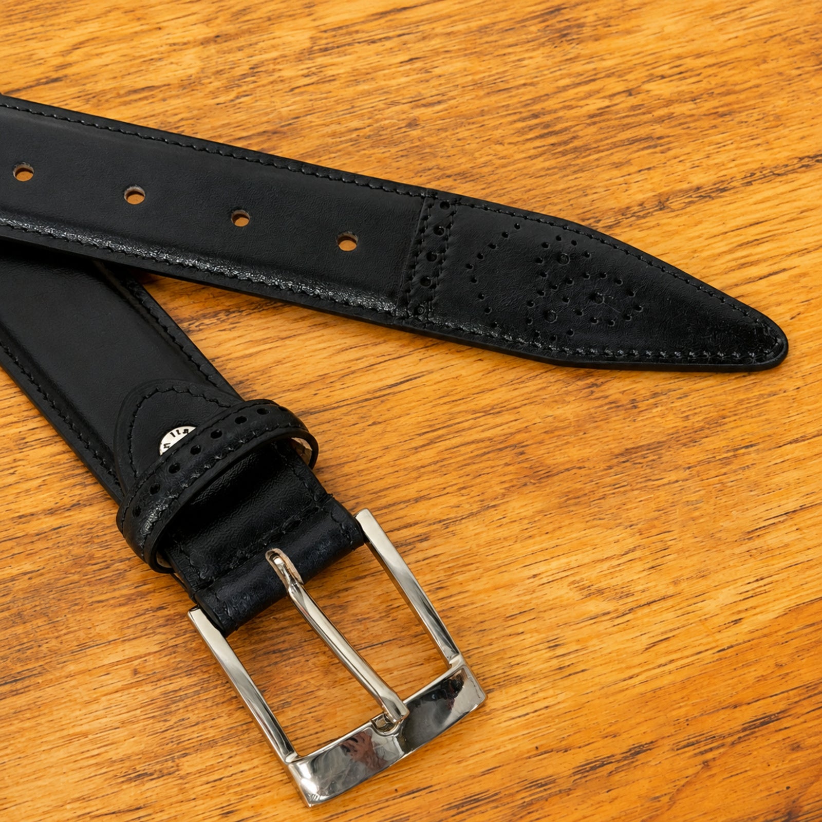 A close-up of a Graphite leather belt with a silver-tone metal buckle, laid diagonally on a warm, textured wooden surface.