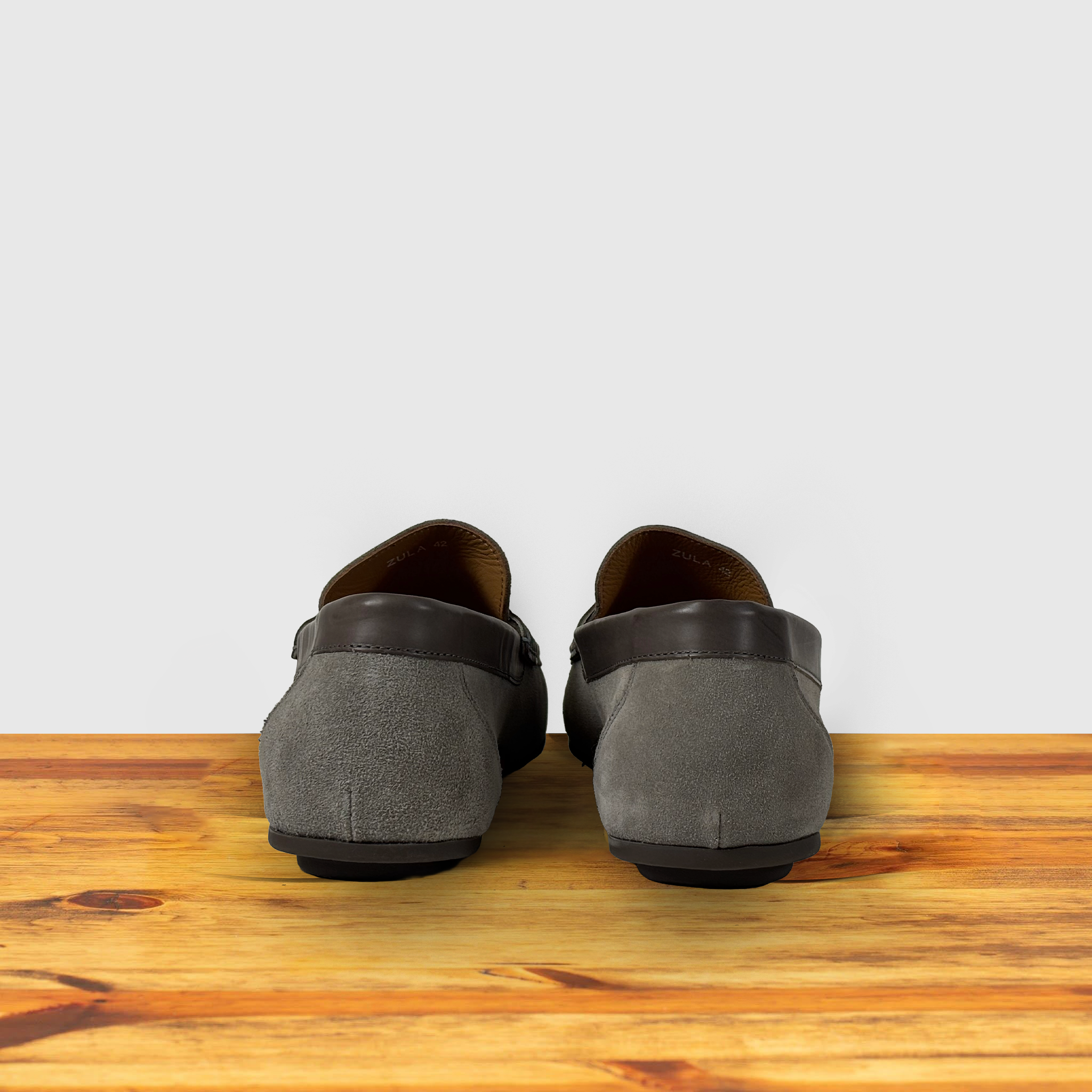 Back side of amsuede penny loafer with a strap, in a gray color, displayed on a wooden surface.