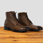 A pair of dark brown textured lace-up ankle boots with a glossy finish, displayed on a wooden surface against a light gray background.
