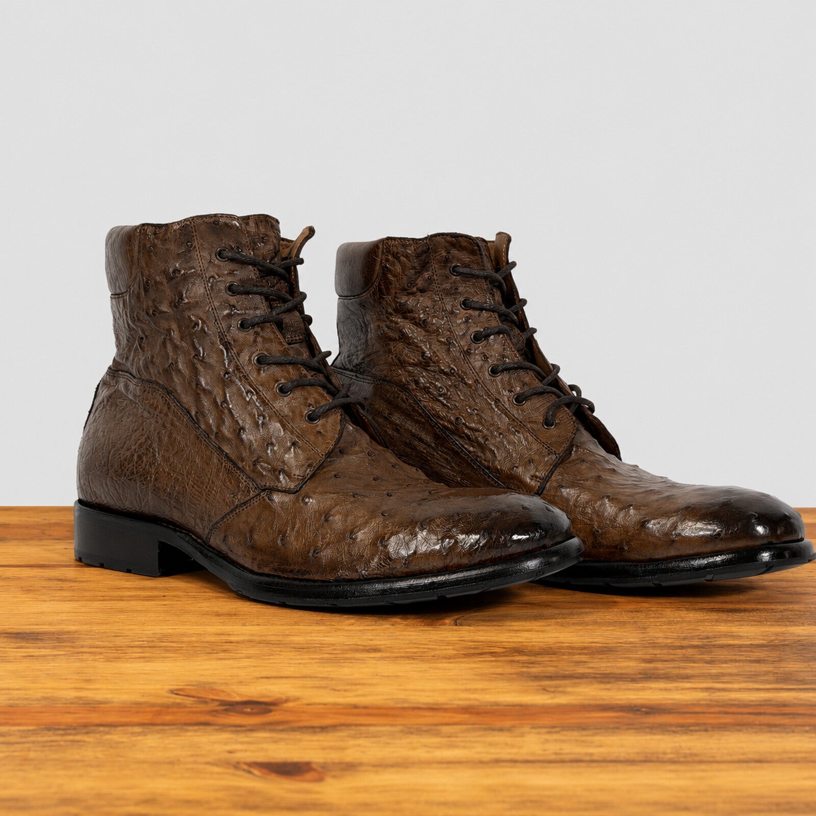 A pair of dark brown textured lace-up ankle boots with a glossy finish, displayed on a wooden surface against a light gray background.