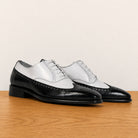 A pair of men's wingtip oxford shoes with a two-tone black and white leather design, displayed on a wooden surface against a plain beige background.
