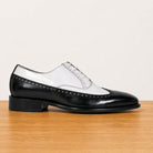 A men's black and white two-tone wingtip oxford shoe with brogue detailing, displayed on a light wood surface against a plain off-white background.
