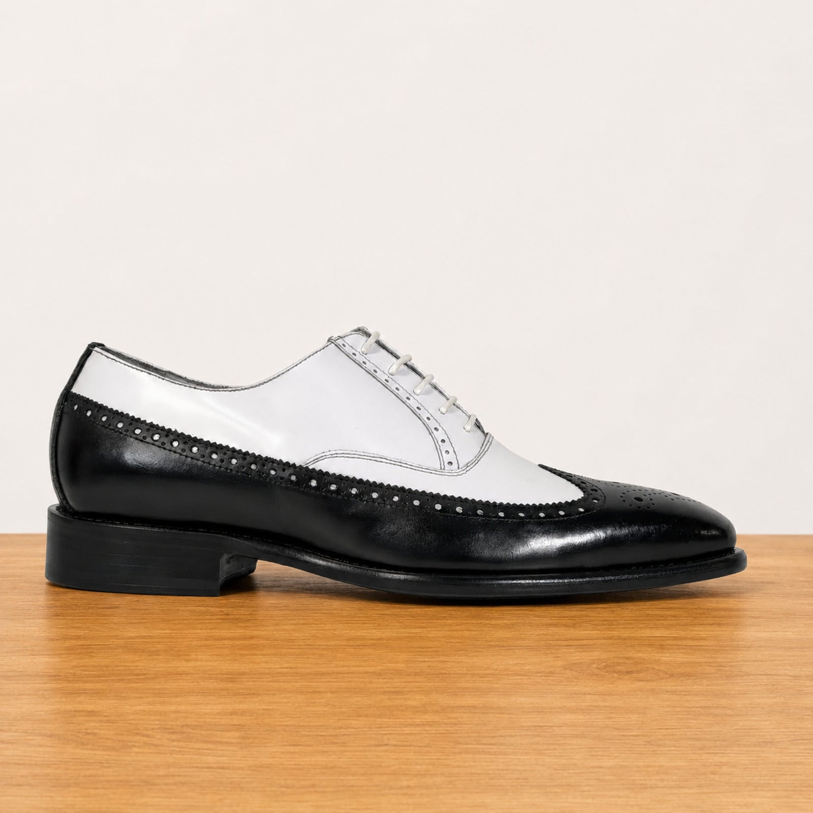 A men's black and white two-tone wingtip oxford shoe with brogue detailing, displayed on a light wood surface against a plain off-white background.