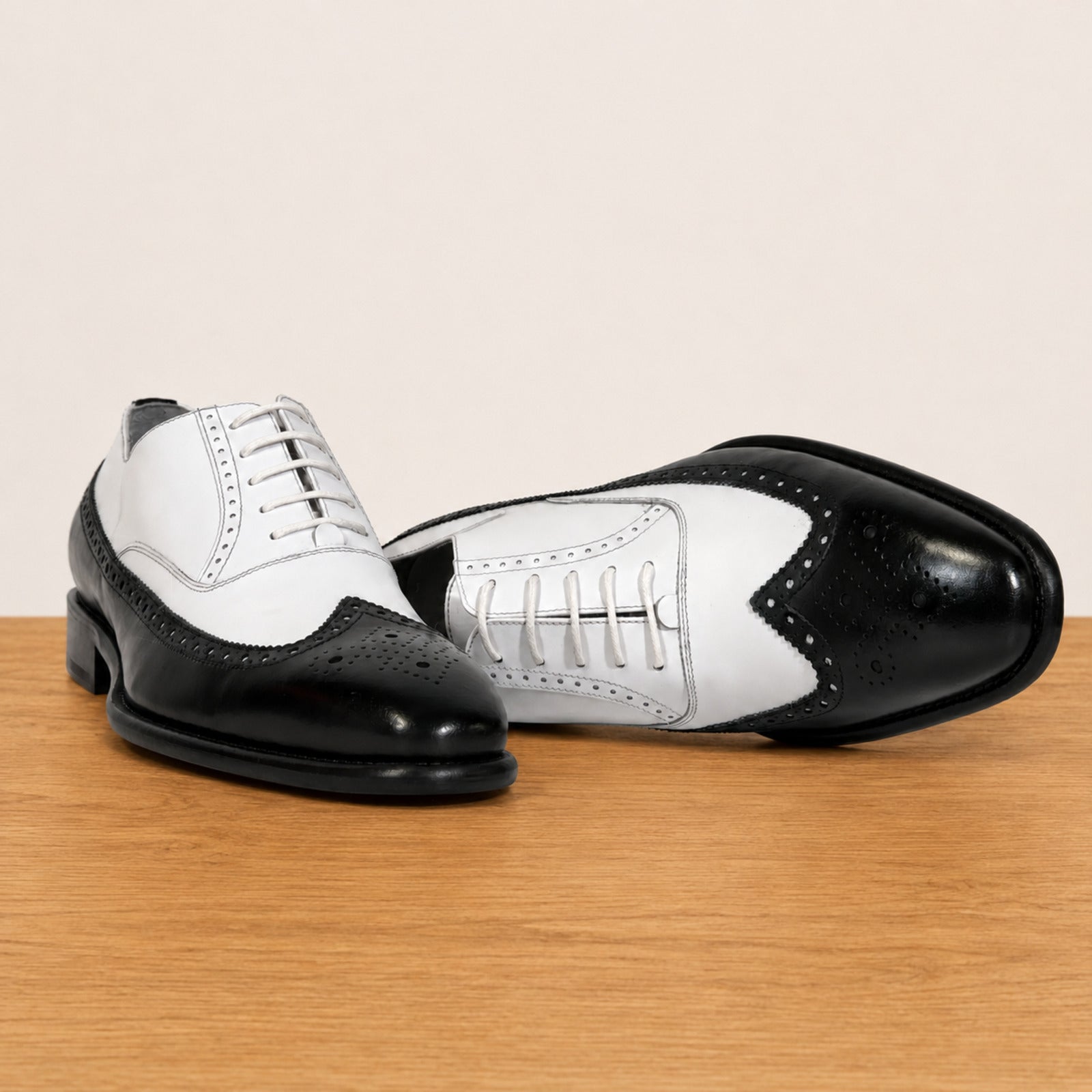 A pair of men's black and white leather wingtip oxford shoes displayed on a light wood surface against a plain beige background.