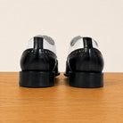 A pair of black and white wingtip oxford shoes viewed from the back, resting on a light brown wooden surface against a plain beige background.