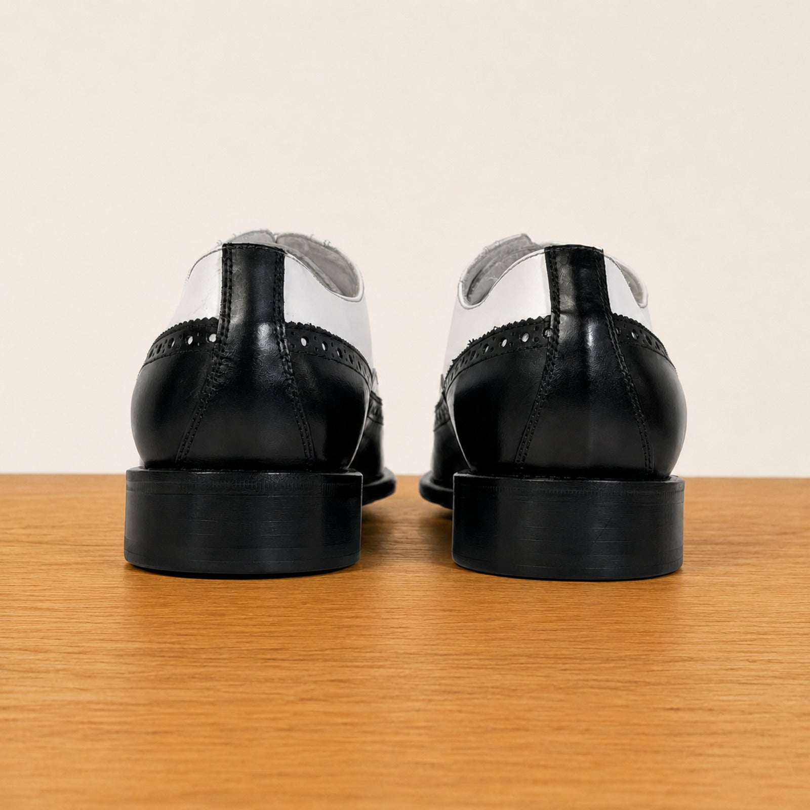 A pair of black and white wingtip oxford shoes viewed from the back, resting on a light brown wooden surface against a plain beige background.