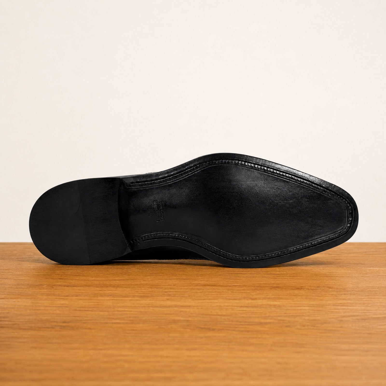 The black leather underside of a dress shoe, showing the outsole and toe cap, resting on a wooden surface against a plain off-white background.